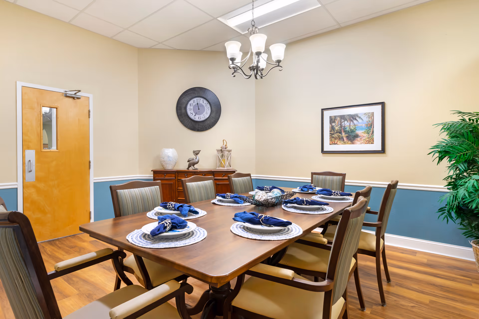 A dining room with a wooden table set for six people with blue napkins on white placemats. The room has beige and blue walls, a wooden door, a wall clock, a framed beach-themed picture, a wooden sideboard with decorative items, a chandelier, and a potted plant in the corner.