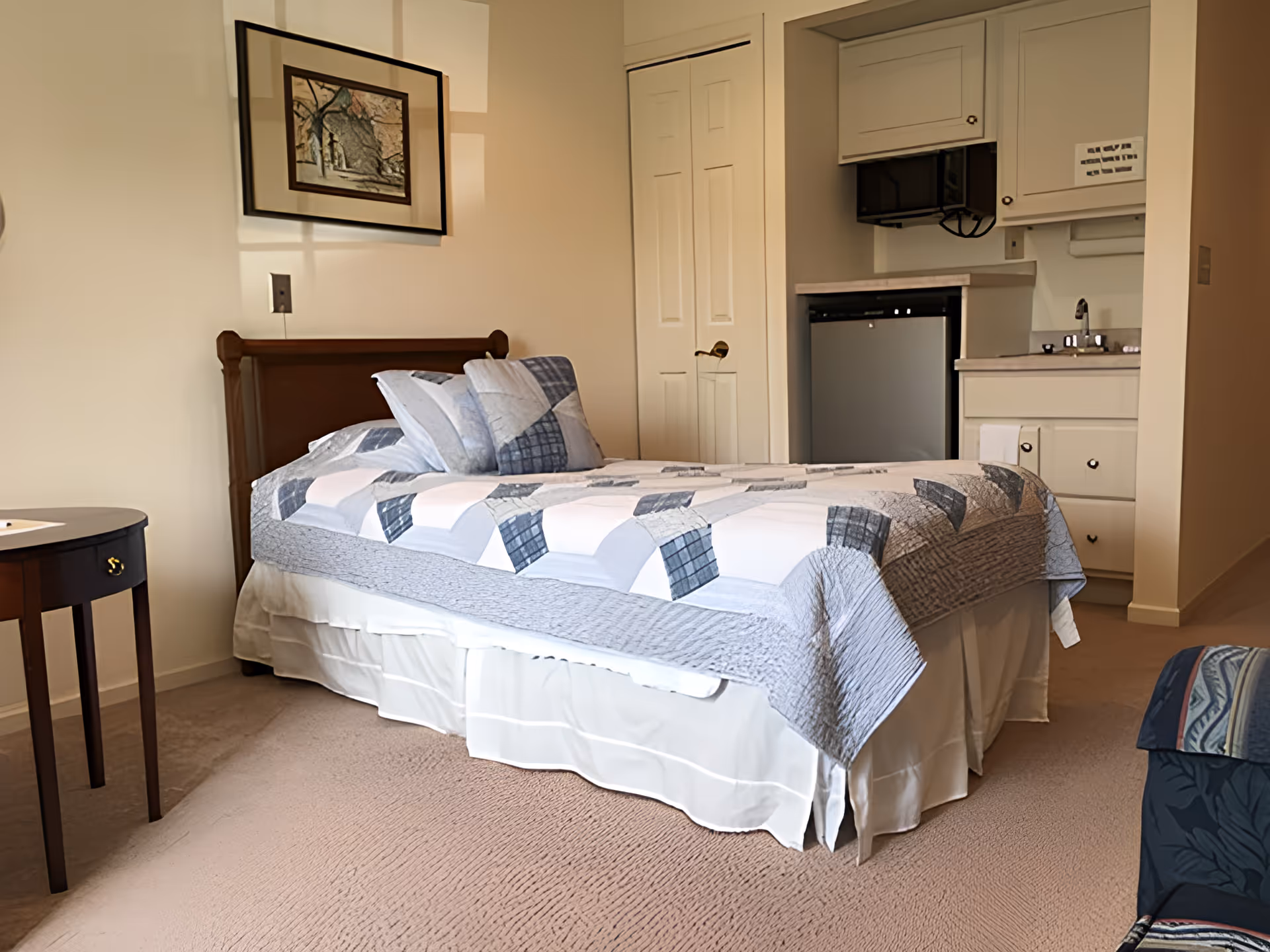 A tidy single bedroom with a made bed, bedside table, wall art, and a small kitchenette with cabinets, mini-fridge, and sink.