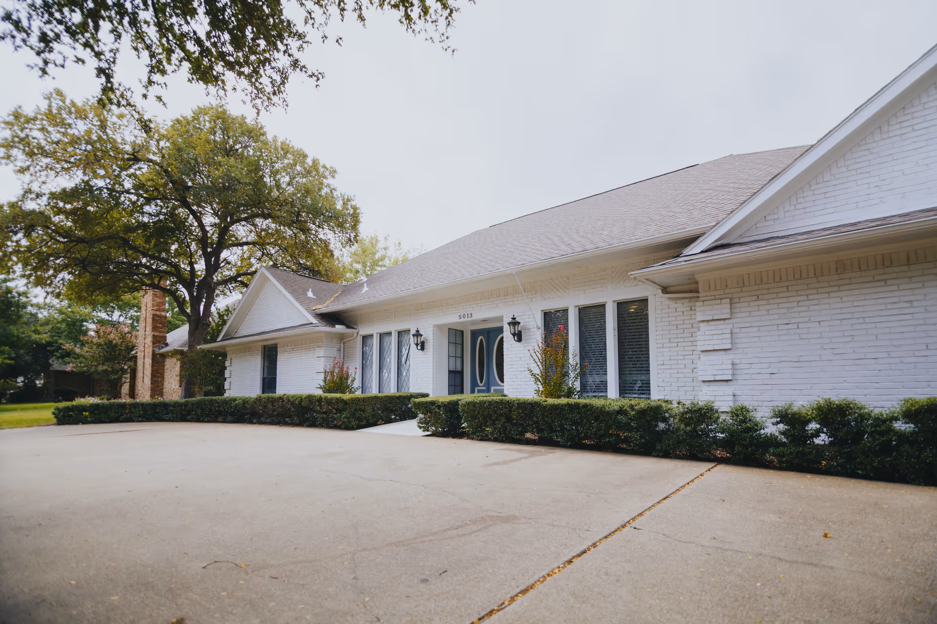 Exterior view of a single-story white brick building with a gray shingled roof, featuring a driveway and trimmed bushes in front. There are large windows and a double door entrance with decorative glass panels. A large tree is visible to the left side of the building.