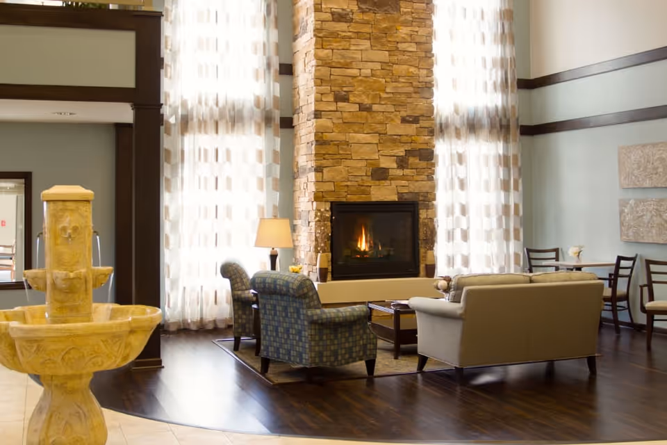 A cozy living room area with a stone fireplace in the center, flanked by tall windows with sheer patterned curtains. The room features a beige sofa, two patterned armchairs, a wooden coffee table, and a floor lamp. To the left, there is a decorative stone water fountain, and in the background, a small dining area with tables and chairs is visible.
