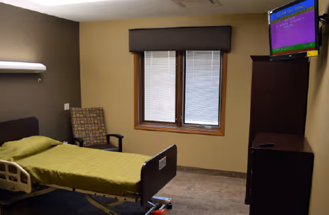 Small resident bedroom with a hospital-style bed, armchair, window with blinds, dresser and wall-mounted TV.