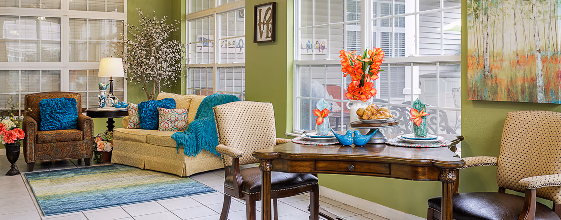 A cozy living room area with a patterned armchair and a beige sofa adorned with blue and patterned cushions. A wooden table is set with plates, napkins, and a vase of bright orange flowers. The room has large windows letting in natural light, green walls, a floor lamp, decorative plants, and colorful artwork on the wall.