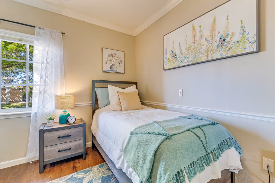 A bright single bedroom with a neatly made bed, nightstand with a lamp and clock, a window with sheer curtains, and artwork on the walls.
