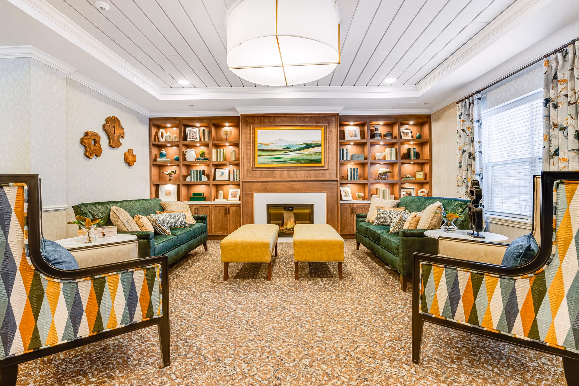 A cozy living room with two green sofas facing each other, two yellow ottomans in the center, and two patterned armchairs with diamond designs in the foreground. The back wall features built-in wooden shelves with decorative items and books, centered around a fireplace with a landscape painting above it. The room has a patterned carpet, floral curtains on the window, and a large ceiling light fixture.