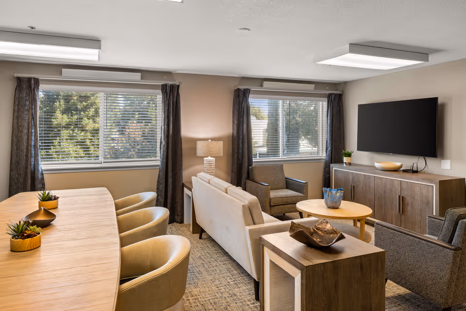 A bright and modern living room in a senior living community featuring a beige sofa, two armchairs, a wooden coffee table, a wooden sideboard with a flat-screen TV mounted above it, and a wooden dining table with four beige chairs. Two large windows with dark curtains allow natural light to fill the room.