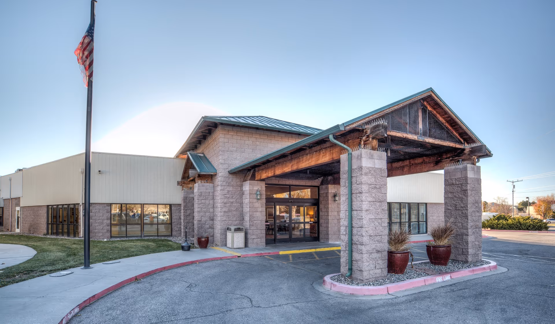 Exterior view of Uptown Rehabilitation Center showing the main entrance with a covered drop-off area supported by stone pillars. There is an American flag on a flagpole to the left and large windows on the building. The sky is clear and the surrounding area includes a parking lot and some landscaping.