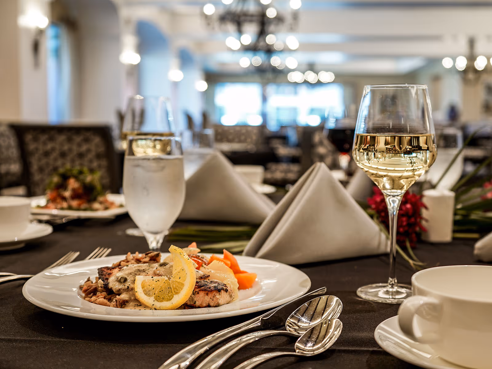 A close-up view of a dining table set with a plate of food garnished with lemon slices, a glass of white wine, a glass of water, folded gray napkins, silverware, and a white coffee cup in a softly lit dining room with upholstered chairs and chandeliers in the background.