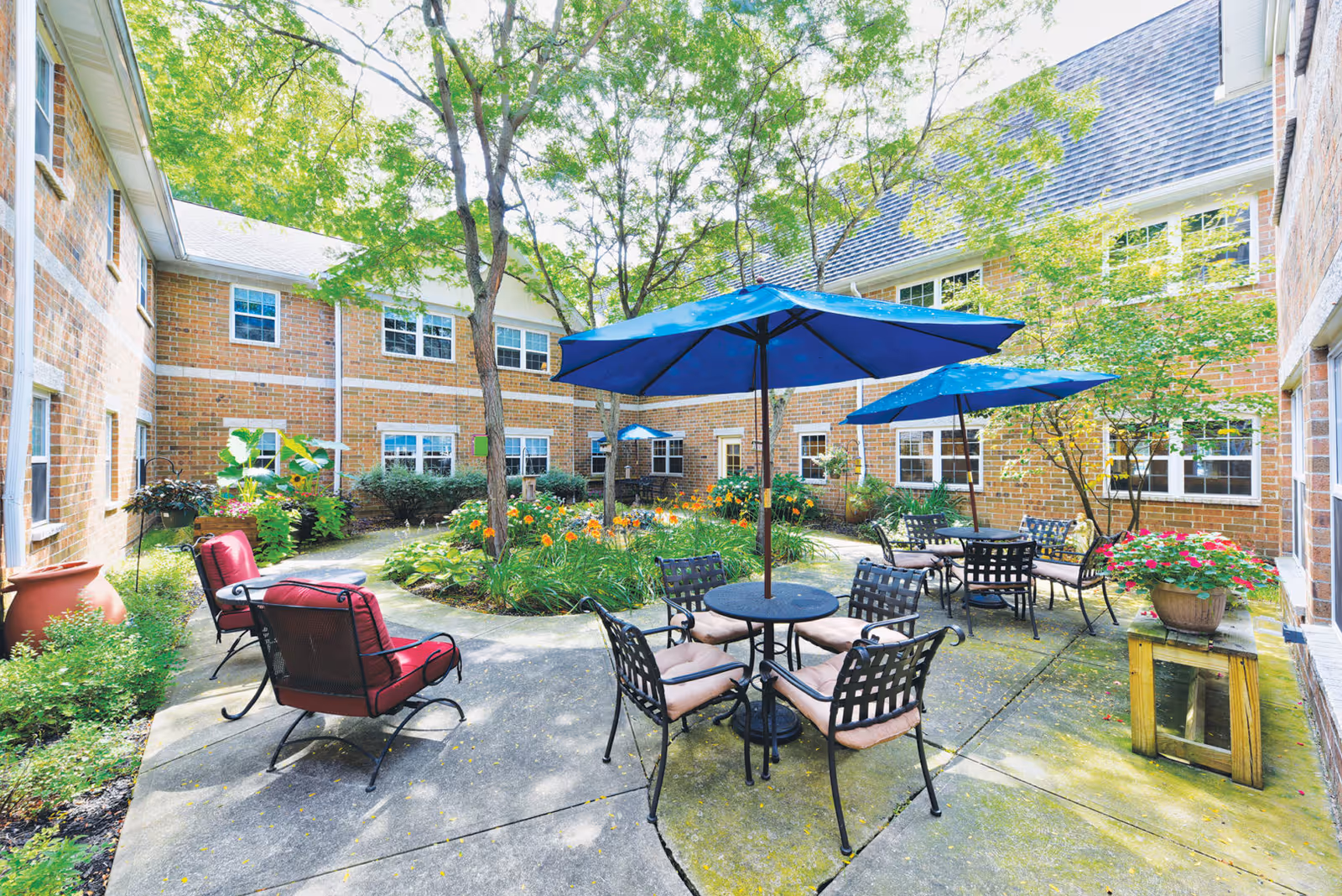 Sunny brick courtyard with patio tables, blue umbrellas, cushioned chairs, and landscaped garden beds between building wings.