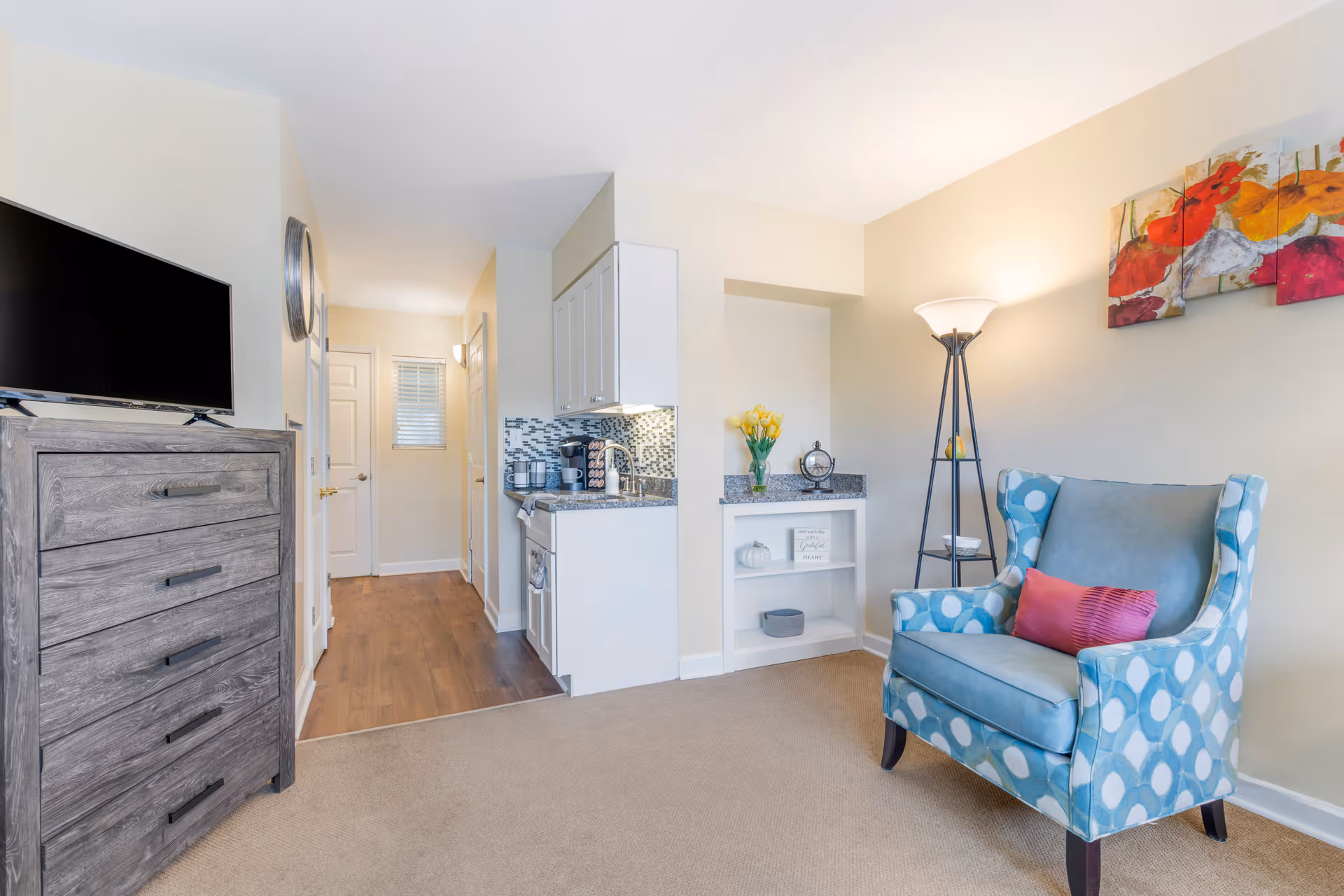 A cozy living area in a senior living facility featuring a blue patterned armchair with a pink pillow, a tall floor lamp, a small built-in shelf with decorative items, and a kitchenette with white cabinets and a coffee maker. A wooden dresser with a flat-screen TV on top is visible on the left, and a hallway with wooden flooring leads to a door with a window above it.