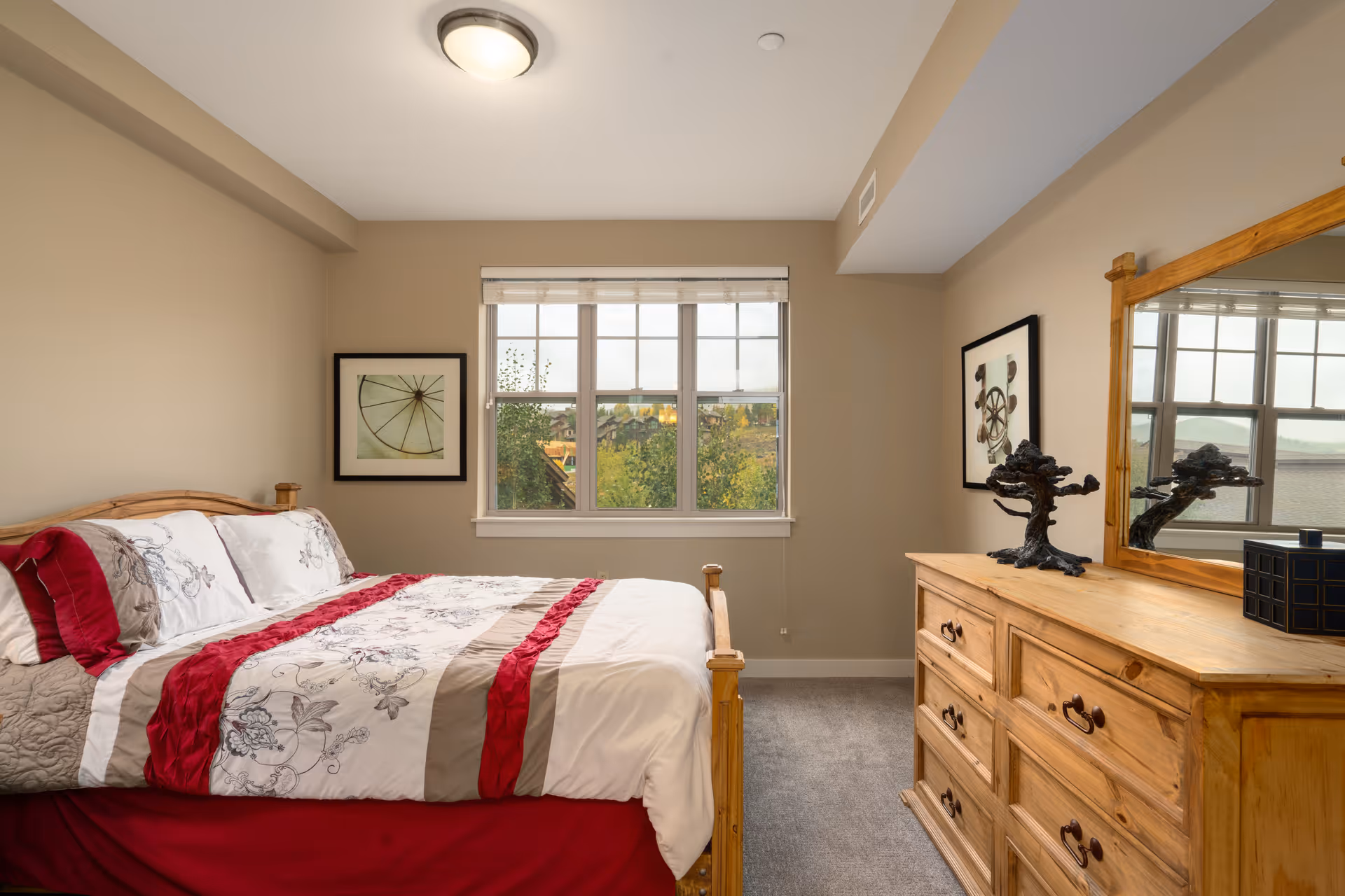 A cozy bedroom with a wooden bed frame and a bed made with white, red, and beige floral bedding. There is a wooden dresser with a large mirror on the right side, decorated with a small tree sculpture and a black box. Two framed pictures hang on the beige walls, and a window in the center shows a view of greenery and houses outside.