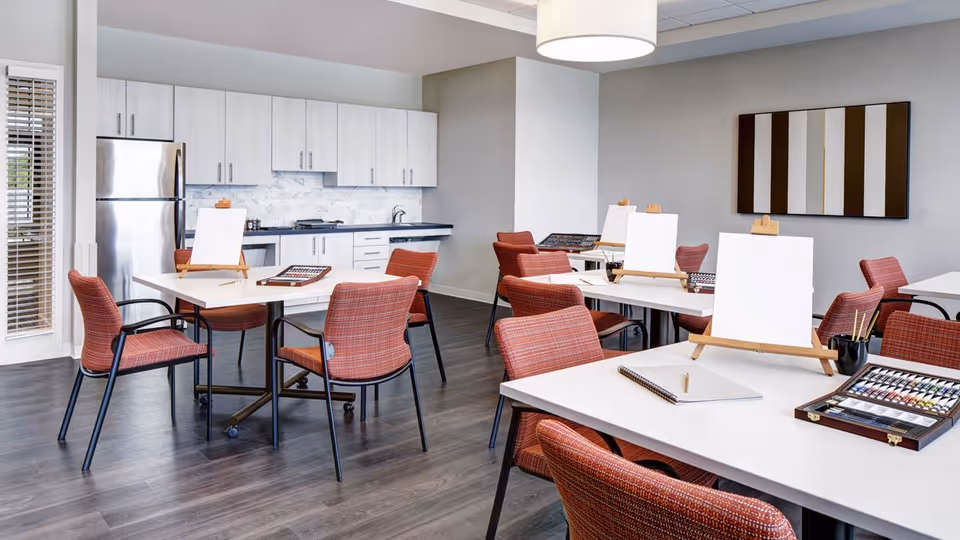 A bright room with multiple tables and red cushioned chairs arranged for an art activity. Each table has a blank canvas on an easel, paint sets, and notebooks. In the background, there is a modern kitchen area with white cabinets, a stainless steel refrigerator, and a marble backsplash. The room has wood flooring and a large light fixture hanging from the ceiling.