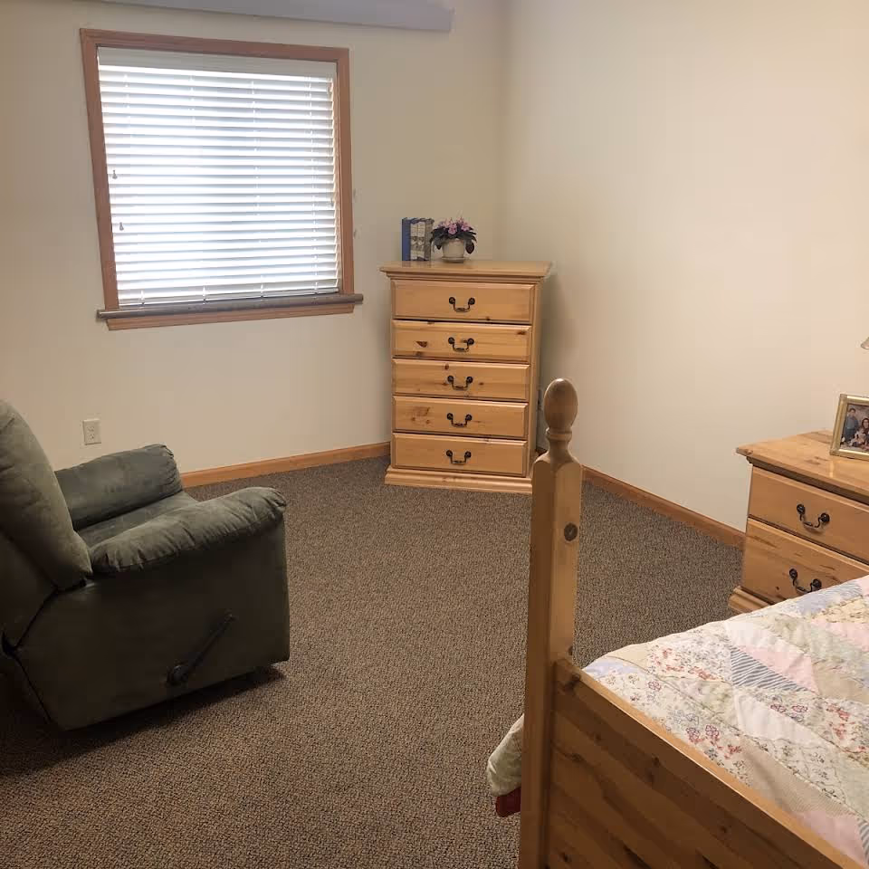 A simple bedroom with a wooden bed partially visible on the right, a green recliner chair on the left, a wooden dresser with five drawers against the far wall, and a window with closed blinds above the dresser. The room has beige walls and carpeted flooring.