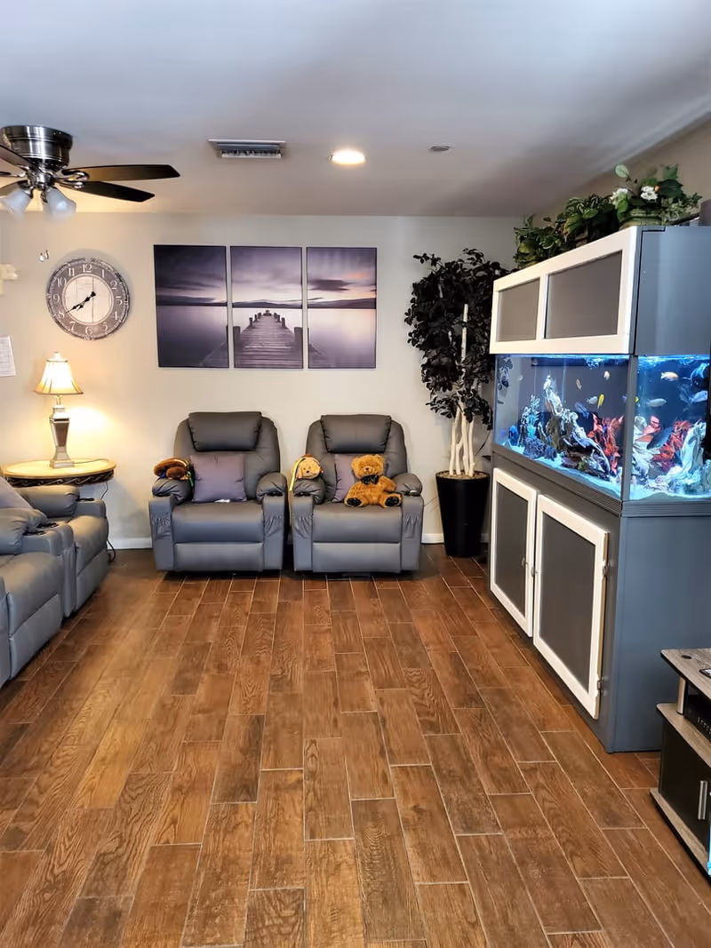A cozy living room with wooden flooring, two gray recliner chairs with teddy bears on them, a large fish tank on the right, a potted plant beside the fish tank, a wall clock, a lamp on a side table, and a three-panel wall art depicting a pier extending into a calm body of water at sunset.