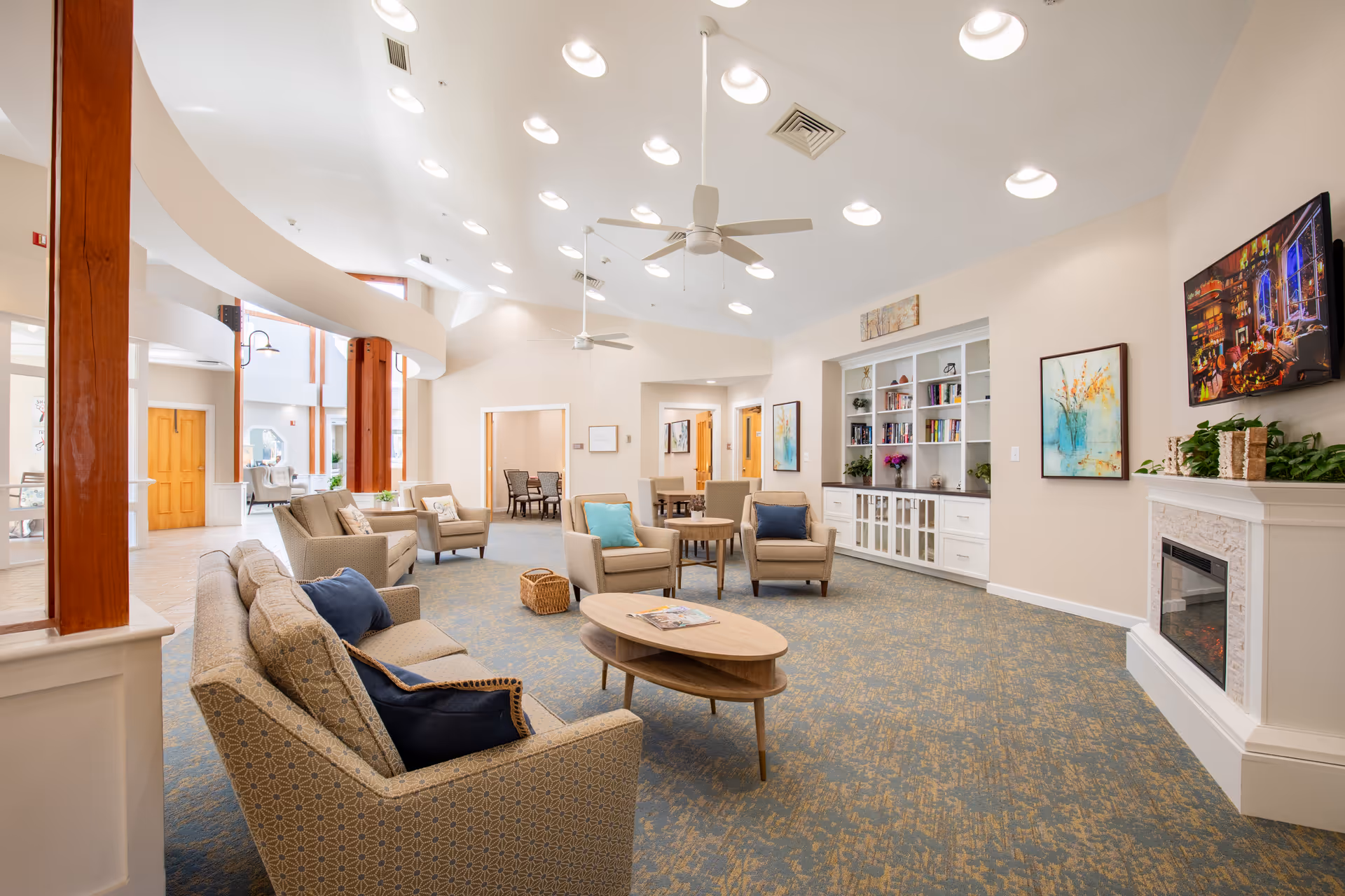 A spacious and well-lit senior living common area with comfortable armchairs and sofas arranged around wooden coffee tables. The room features a built-in bookshelf, a wall-mounted TV above a white fireplace, ceiling fans, and decorative artwork on the walls. Large windows and high ceilings with recessed lighting create an inviting atmosphere.