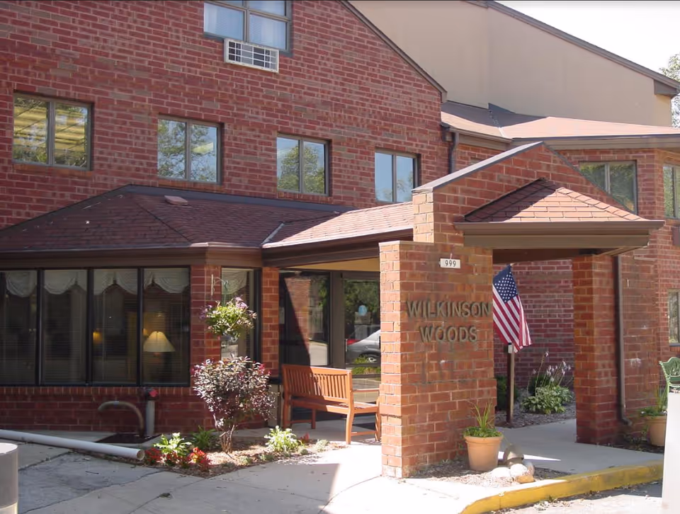 Brick front entrance of Wilkinson Woods Senior Community showing a covered portico with a bench, potted plants, windows, and an American flag.