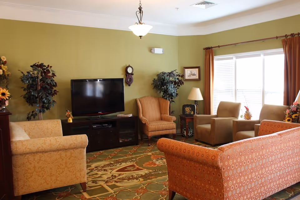A cozy living room in Meadow View Senior Living with a green wall, a flat-screen TV on a dark wooden stand, an orange patterned sofa, a beige armchair, two brown armchairs, a striped armchair, a side table with a lamp, potted plants, a wall clock, and a large window with orange curtains letting in natural light.