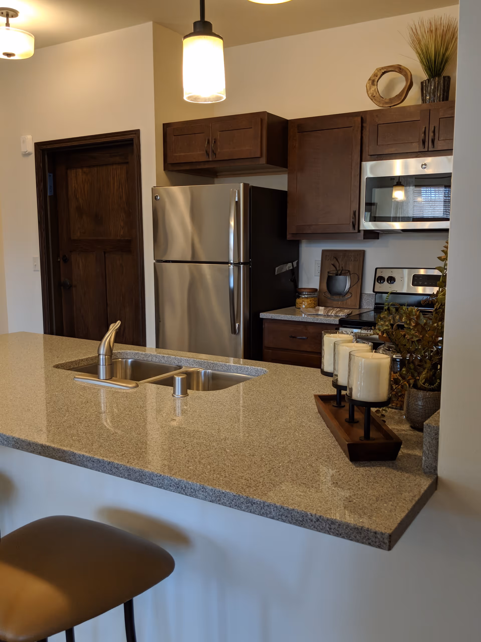 Modern kitchen with a granite countertop island featuring a built-in sink and faucet, a stainless steel refrigerator, stove, and microwave. Dark wood cabinets are mounted on the walls, and decorative items including candles and plants are placed on the countertop. A brown cushioned bar stool is partially visible in the foreground.
