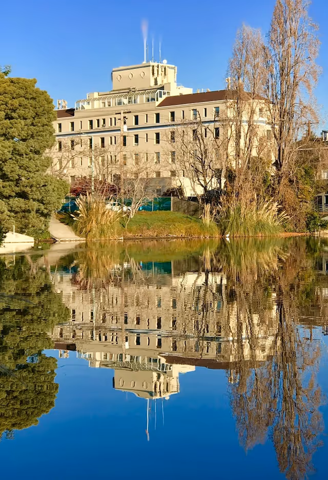 A large multi-story beige building reflected clearly in a calm body of water, surrounded by trees and greenery under a clear blue sky.