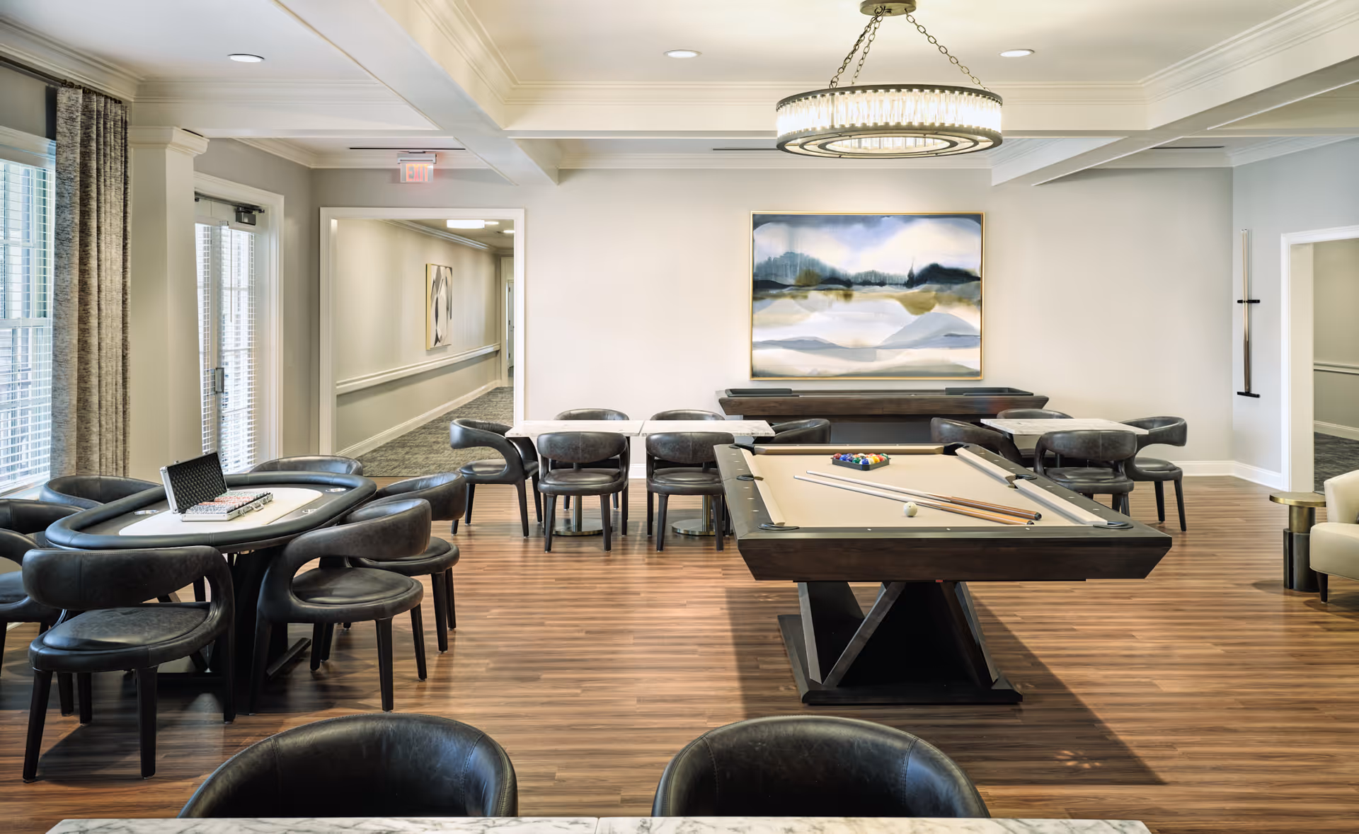 Bright communal lounge with a pool table centered on wood floors, surrounded by round game tables, chairs, and wall artwork.