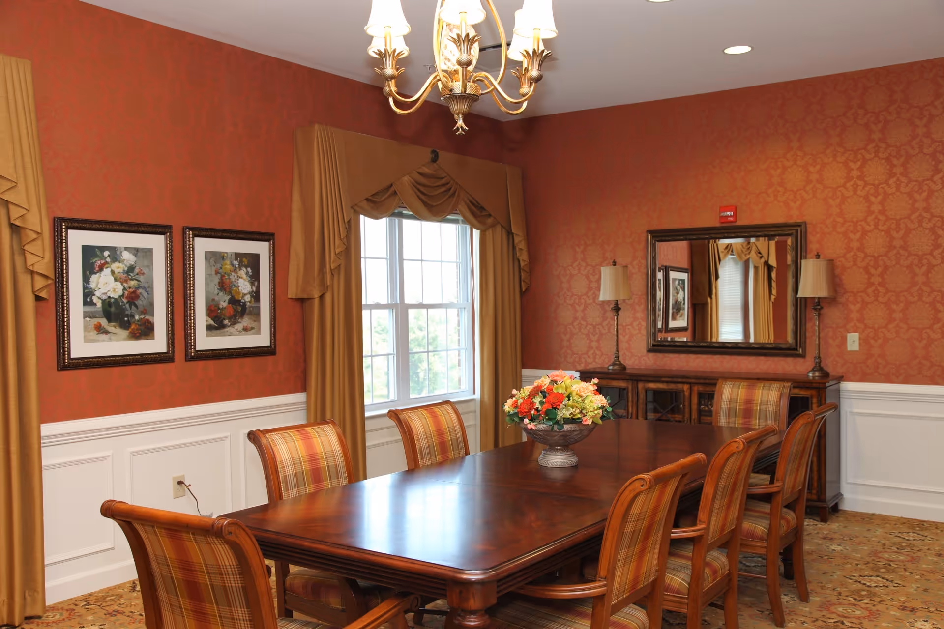 A formal dining room with a long wooden table surrounded by six plaid upholstered chairs. The walls are decorated with red patterned wallpaper and white wainscoting. Two framed floral paintings hang on one wall, and a large window with gold drapes lets in natural light. A chandelier hangs above the table, and a sideboard with two lamps and a large mirror is against the far wall. A floral centerpiece is on the table.