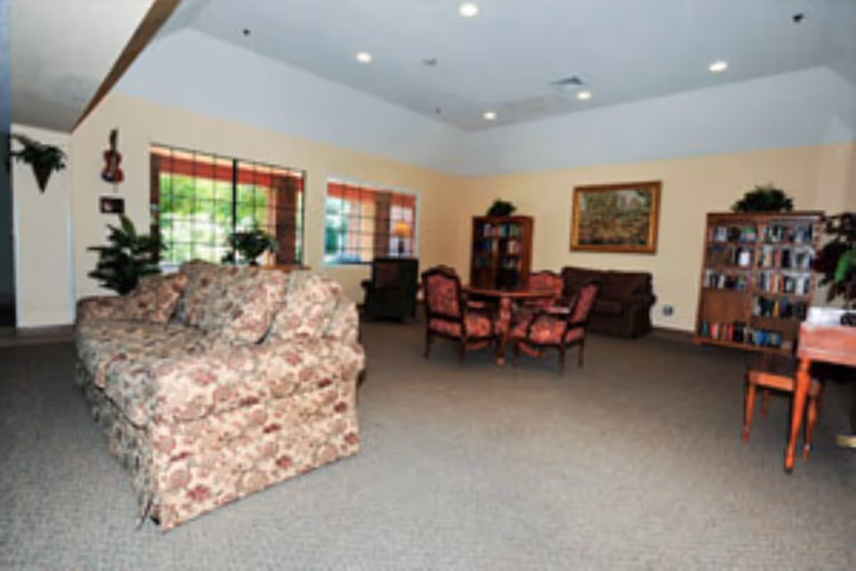 A spacious common area with floral patterned sofas, a round table with four upholstered chairs, bookshelves filled with books, potted plants, and a framed painting on the wall. Large windows allow natural light to enter the room.