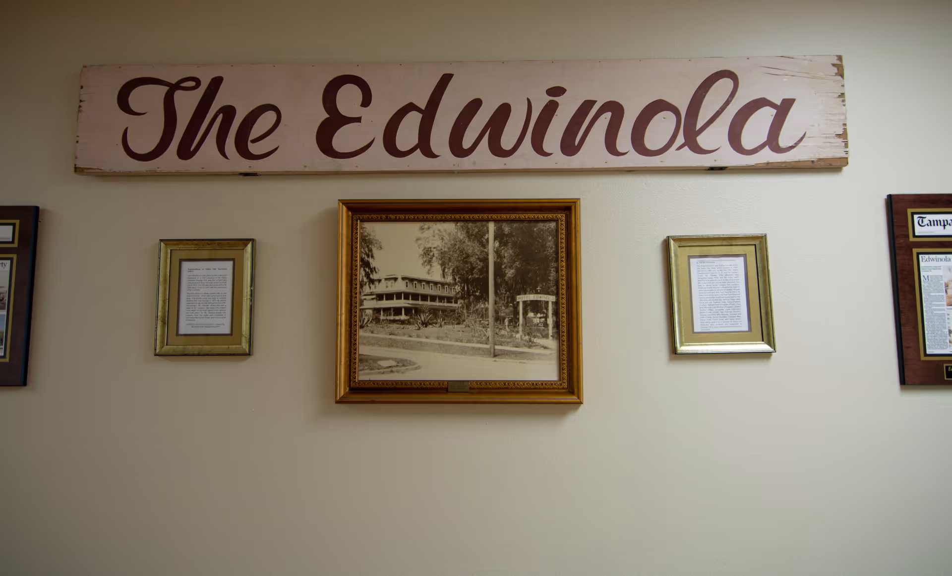 A wall with a wooden sign reading 'The Edwinola' above framed pictures and documents, including a sepia-toned photograph of a building surrounded by trees.