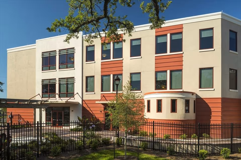 Three-story beige and terracotta modern building façade with rows of windows, a fenced landscaped front and entry canopy.