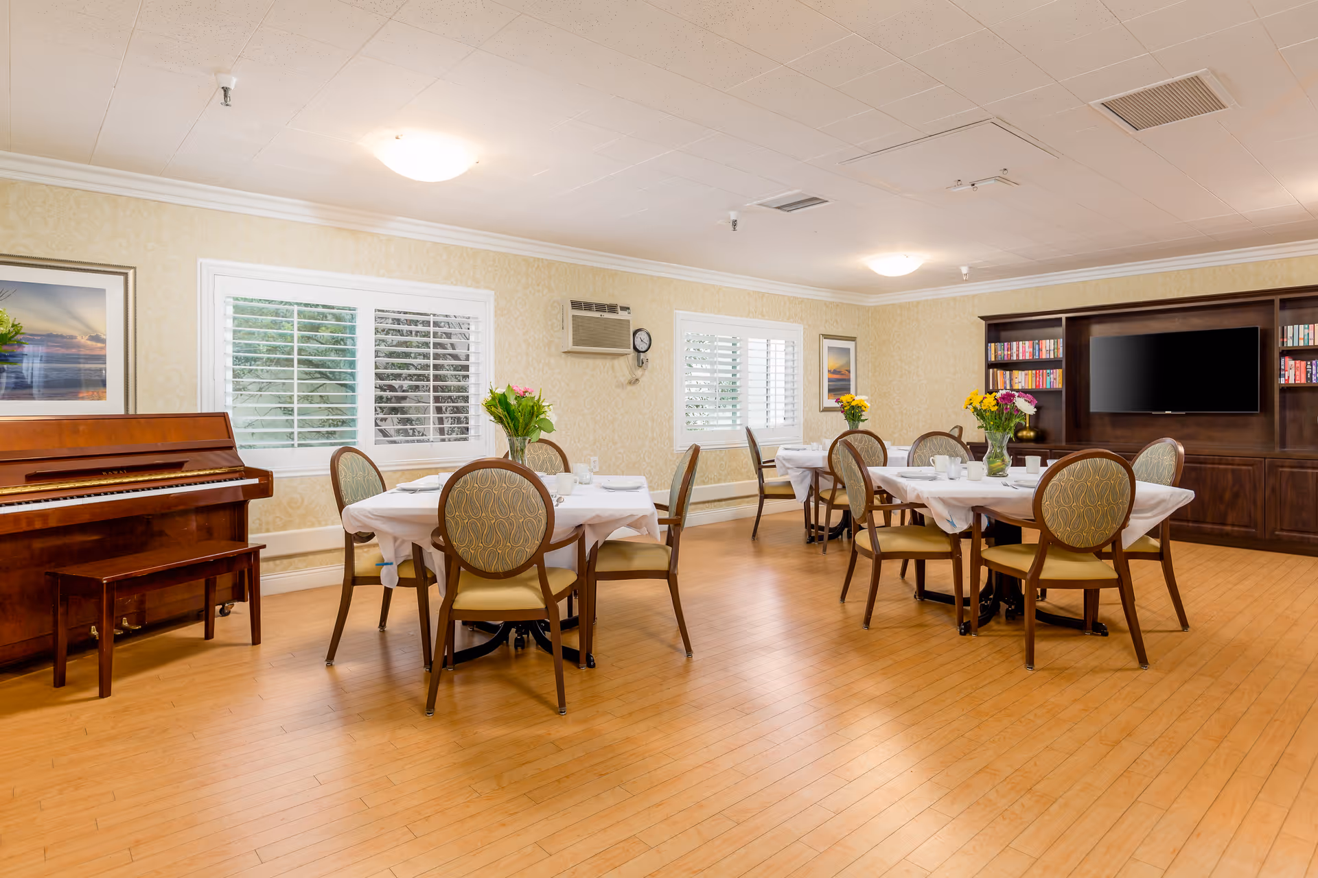 A bright dining room with wooden floors, round tables covered with white tablecloths, and chairs arranged around them. Each table has a vase with colorful flowers. There is a wooden piano on the left side and a large wooden bookshelf with a flat-screen TV on the right wall. The room has two windows with white shutters and framed pictures on the walls.