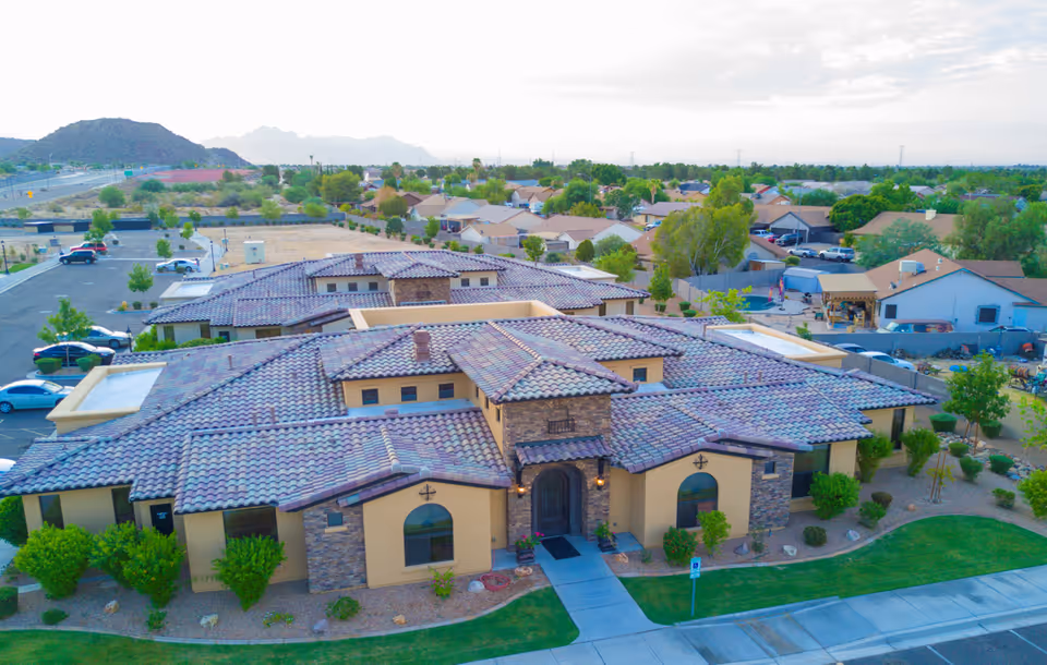 Aerial view of a single-story senior living facility building with a tiled roof and stone accents, surrounded by landscaped greenery and a parking lot. Residential houses and mountains are visible in the background under a partly cloudy sky.