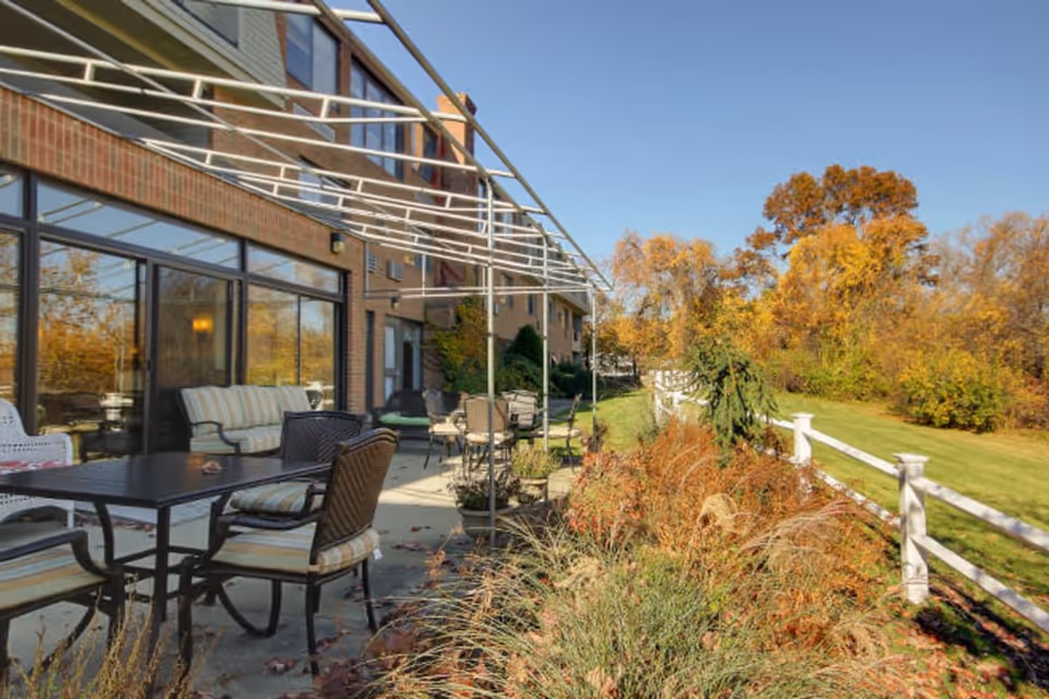 Outdoor patio area at a senior living facility with tables and cushioned chairs arranged along a concrete walkway next to a building with large windows. There is a white fence bordering a grassy area with autumn-colored trees and shrubs under a clear blue sky.