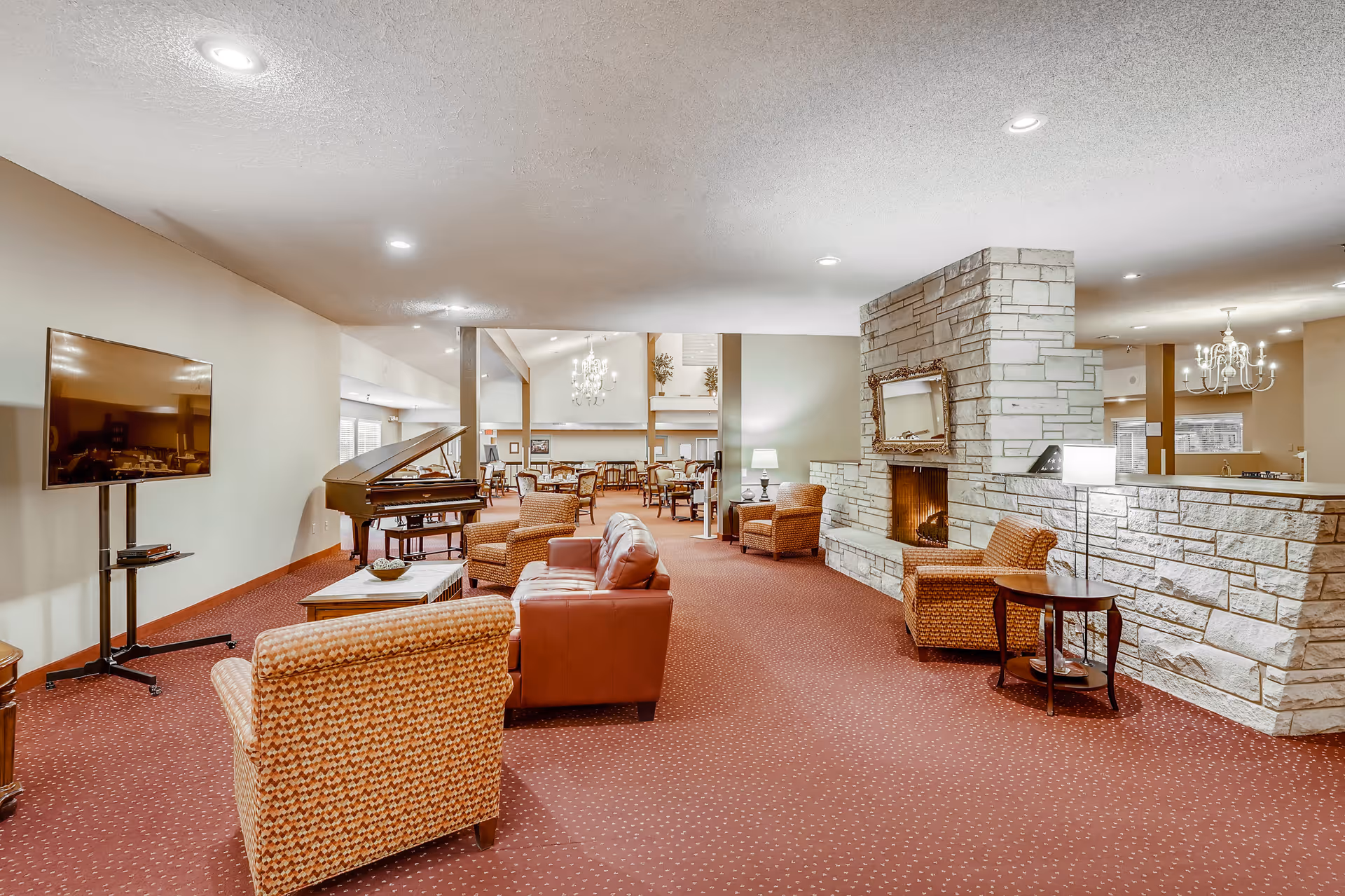 A spacious living room area in a senior living facility featuring a red carpet, multiple armchairs and sofas, a flat-screen TV mounted on a stand, a grand piano, and a large stone fireplace with a decorative mirror above it. In the background, there is a dining area with tables and chairs, and chandeliers hanging from the ceiling.