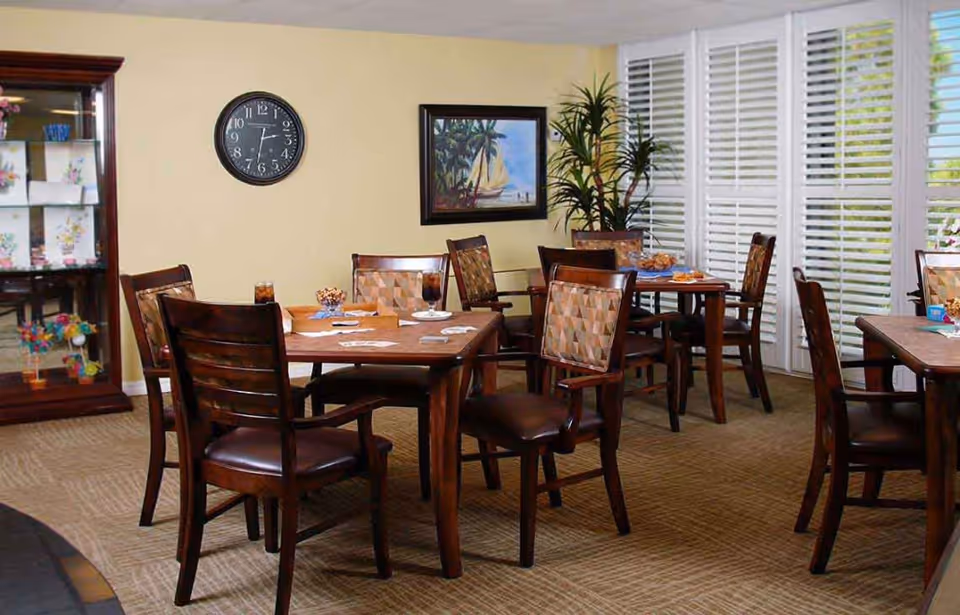 A cozy dining room with several wooden tables and chairs arranged neatly. The tables have drinks and snacks on them. The room has beige walls, a clock, a framed painting of a sailboat, a tall plant, and large windows with white blinds letting in natural light. A display cabinet with decorative items is visible on the left side.