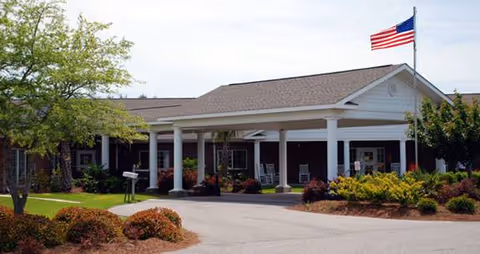 Exterior view of a single-story building with a covered entrance supported by white columns. There is an American flag flying on a flagpole above the entrance. The surrounding area features landscaped bushes, small trees, and a paved driveway leading to the entrance.
