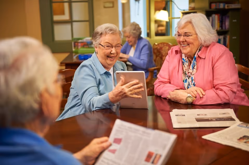 Three elderly women sitting around a wooden table in a cozy room. One woman in a light blue jacket is holding a tablet and smiling, another woman in a pink jacket with a scarf is also smiling, and a third woman is holding a newspaper. In the background, another elderly woman is seated and engaged in an activity. The room has warm lighting and bookshelves.