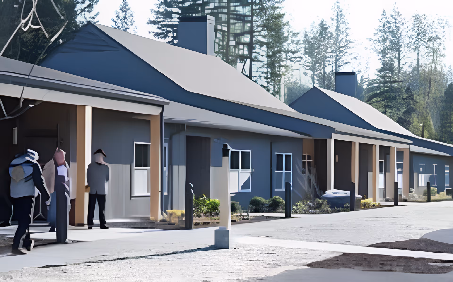 Exterior view of a single-story building with a covered walkway and several windows, surrounded by trees and greenery. There are a few people walking near the entrance.