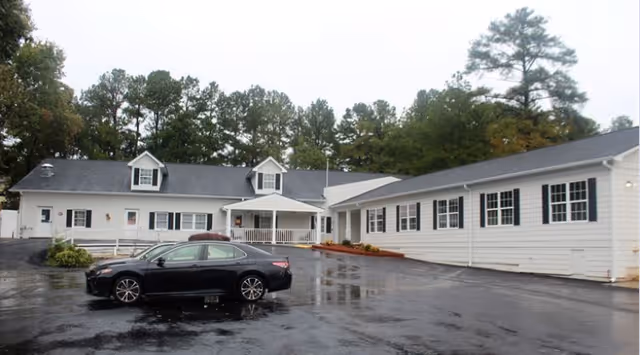 Exterior view of a single-story white building with black shutters and a dark roof, surrounded by trees. A black car is parked on the wet asphalt driveway in front of the building.