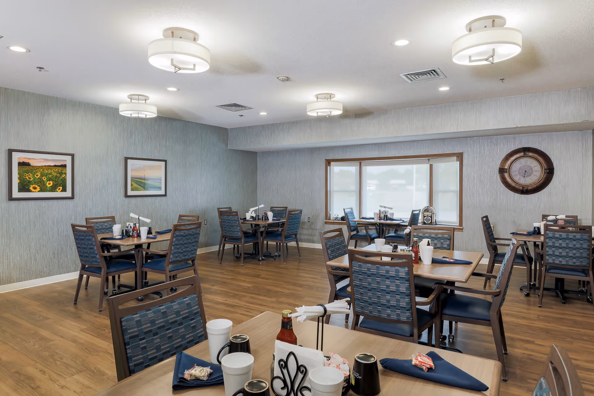 A dining room in an assisted living facility with multiple tables and chairs arranged neatly. Each table has place settings including napkins, cups, and condiments. The room has wooden flooring, light gray textured walls, framed pictures on the wall, a large clock, and ceiling lights providing bright illumination. A window with blinds is visible in the background.