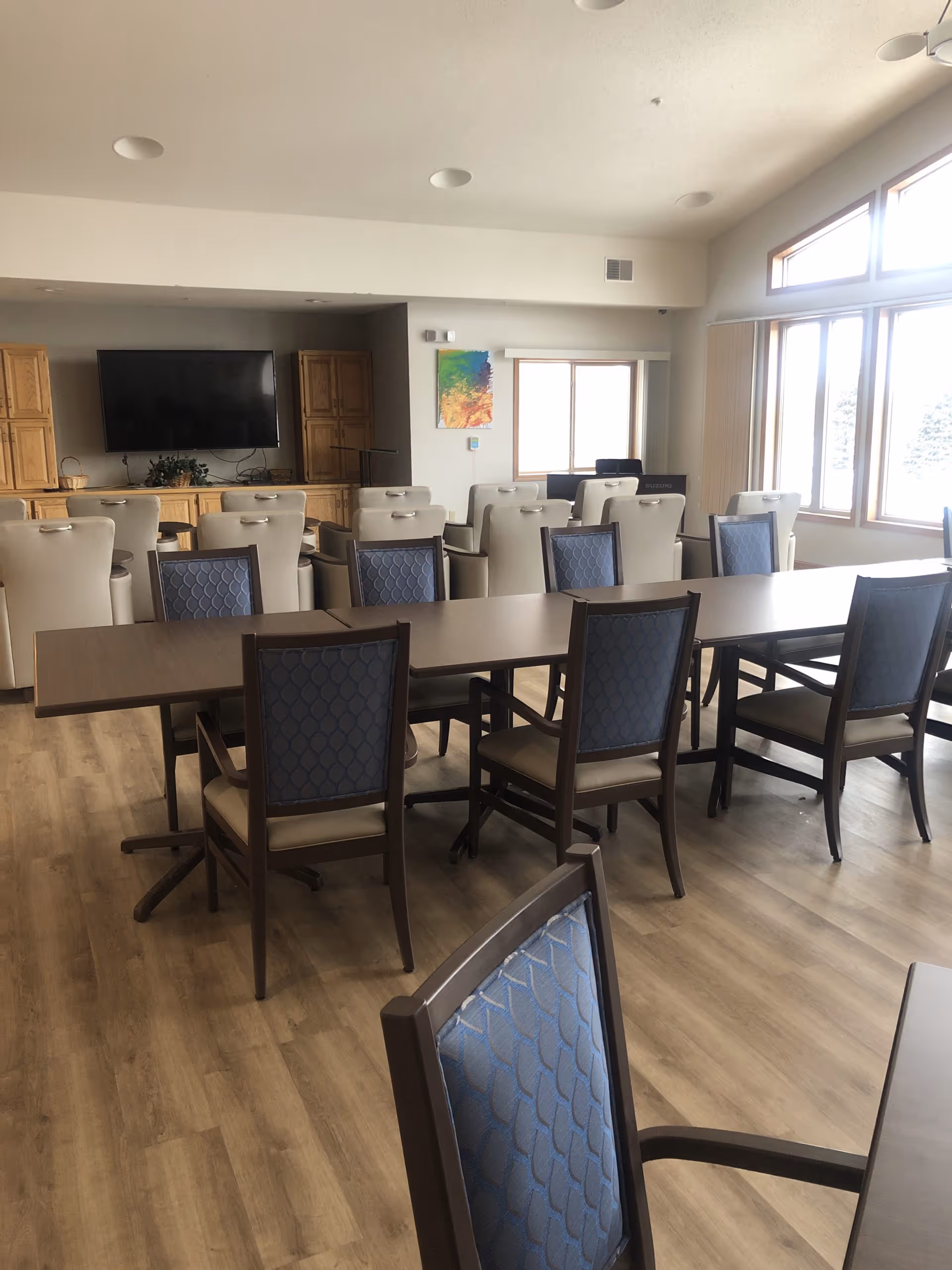 A senior living facility common area with multiple tables and chairs arranged for group seating. The room has large windows letting in natural light, a wall-mounted flat-screen TV, wooden cabinets, and a colorful abstract painting on the wall. The flooring is wood-style laminate.
