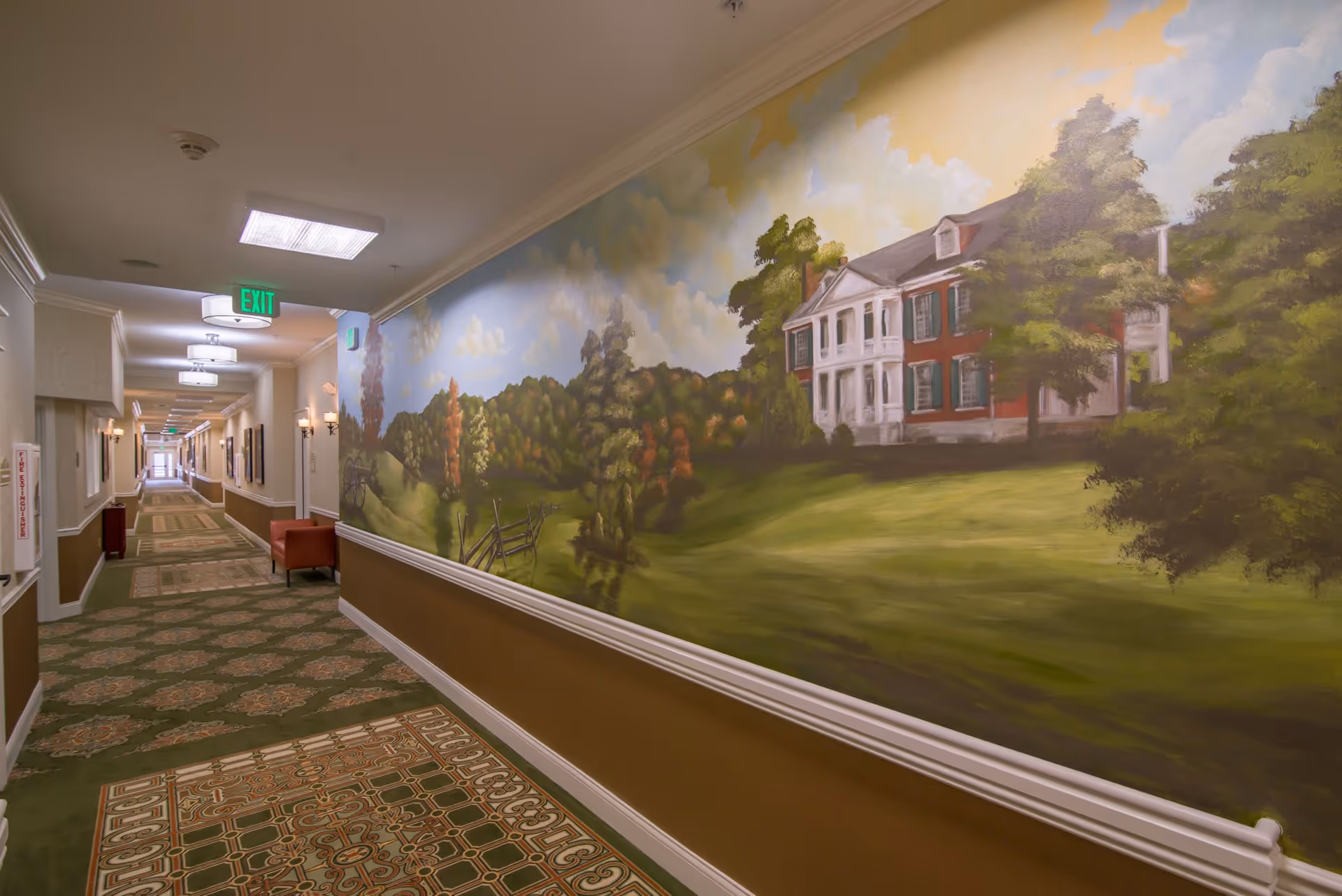 A long indoor hallway with patterned carpet and beige walls. The right wall features a large mural painting depicting a countryside scene with a large house, trees, and a fence under a partly cloudy sky. The hallway is lit with ceiling lights and wall sconces, and there is an exit sign visible at the far end.