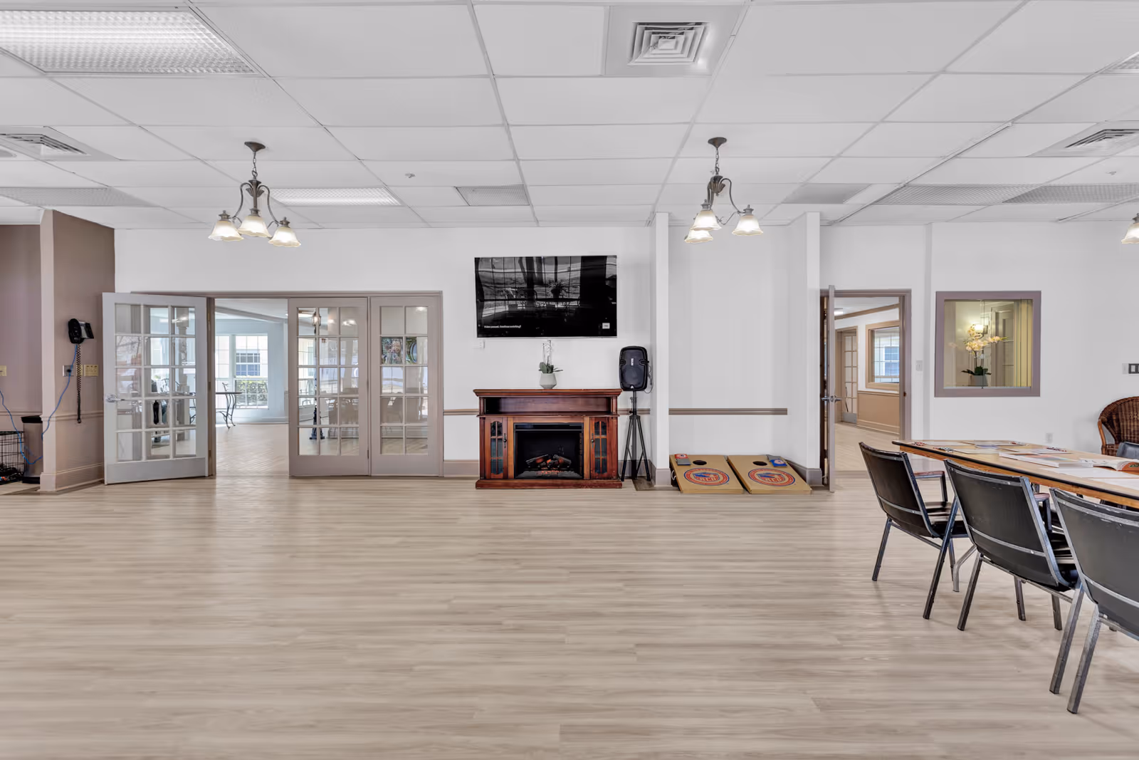 A spacious common area in an assisted living facility with light wood flooring, white walls, and a ceiling with recessed lighting and hanging lamps. There is a flat-screen TV mounted above a wooden electric fireplace. To the right of the fireplace are two cornhole boards leaning against the wall. On the right side of the image, there is a table with several chairs around it. French doors and open doorways lead to other rooms with large windows allowing natural light to enter.