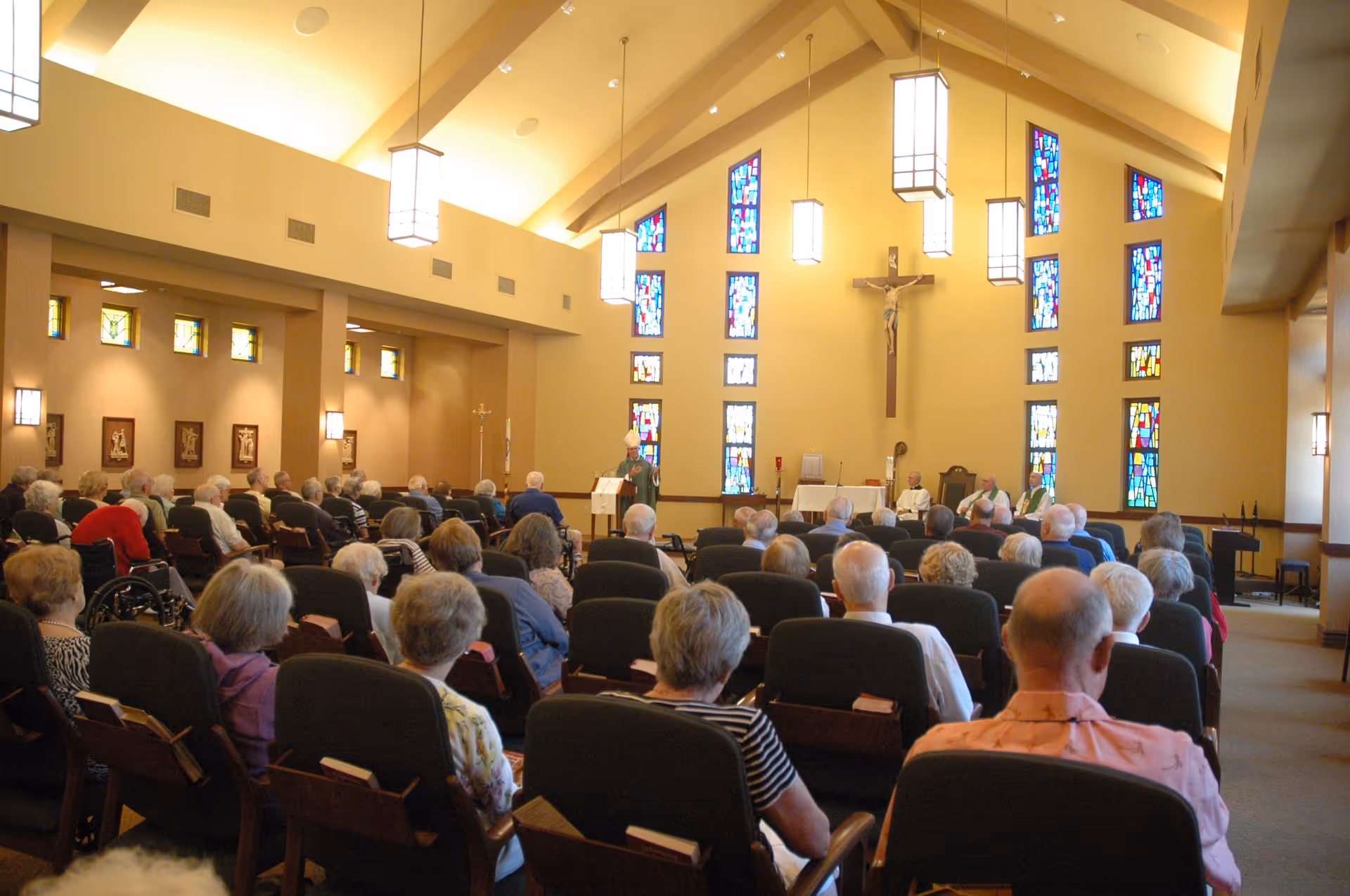 Interior view of a chapel with stained glass windows and a crucifix on the wall. Several elderly people are seated in rows of chairs facing the altar where clergy members are conducting a service.