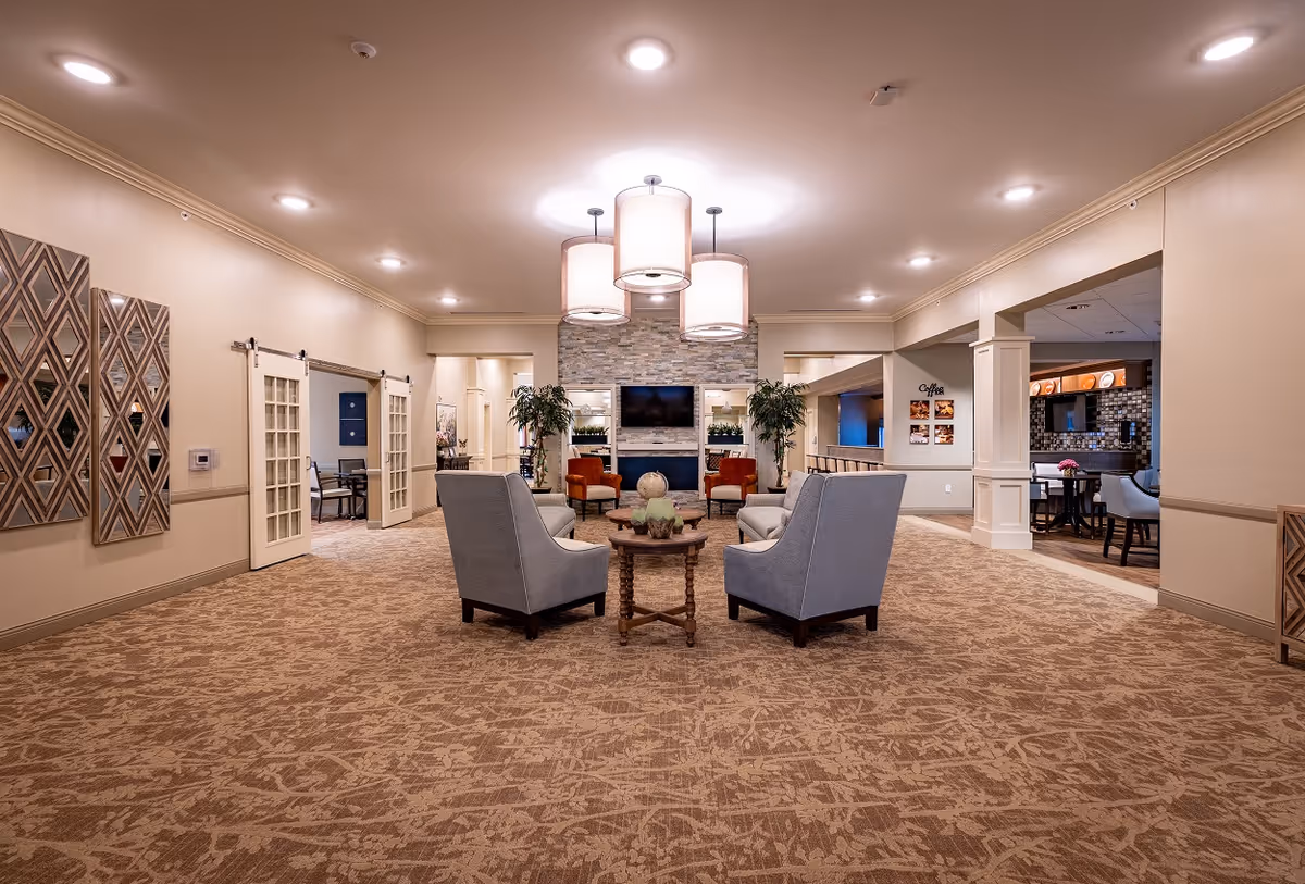Spacious communal living room with armchairs arranged around a central table, a fireplace and TV at the far wall, and pendant lights overhead.