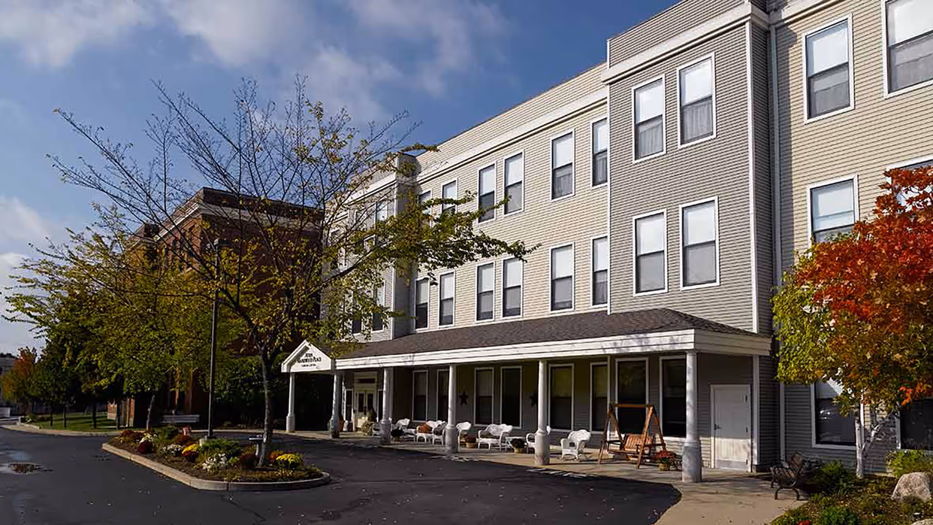 Exterior view of a three-story senior living facility building with beige and gray siding, multiple windows, a covered porch with white columns, outdoor seating, and trees with autumn foliage along the driveway.