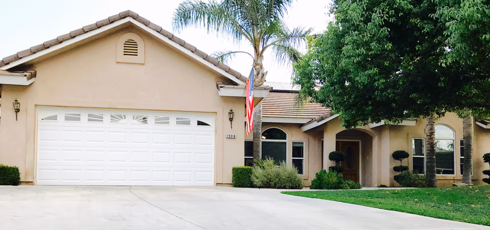Front exterior view of a single-story house with a beige facade, a white garage door, an American flag mounted on a pole near the entrance, and well-maintained landscaping including palm trees and bushes.
