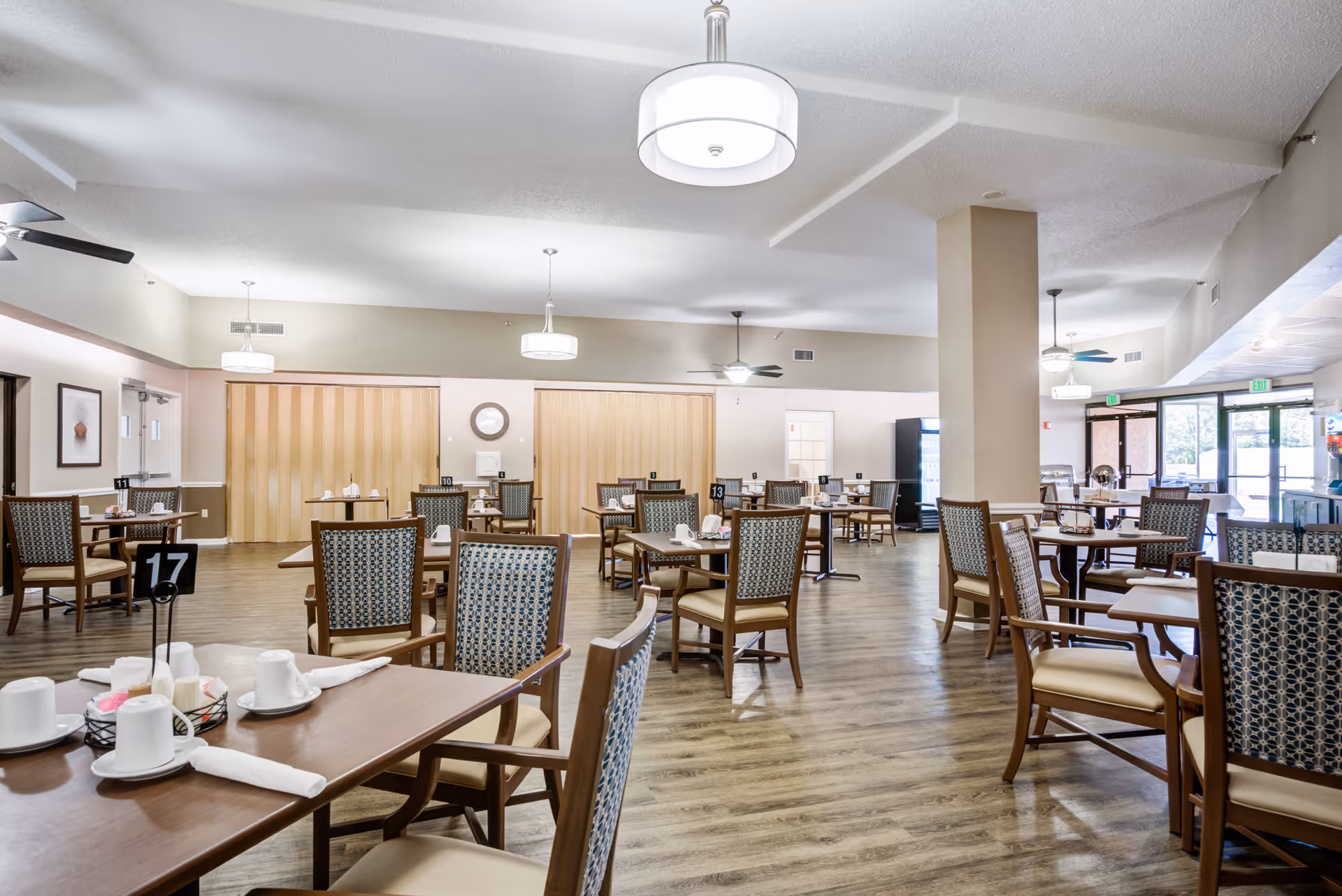 A spacious dining room with multiple wooden tables and cushioned chairs arranged neatly. Each table is set with white cups, napkins, and condiments. The room has wooden flooring, ceiling fans, and modern hanging light fixtures. Large windows and glass doors allow natural light to enter the space.