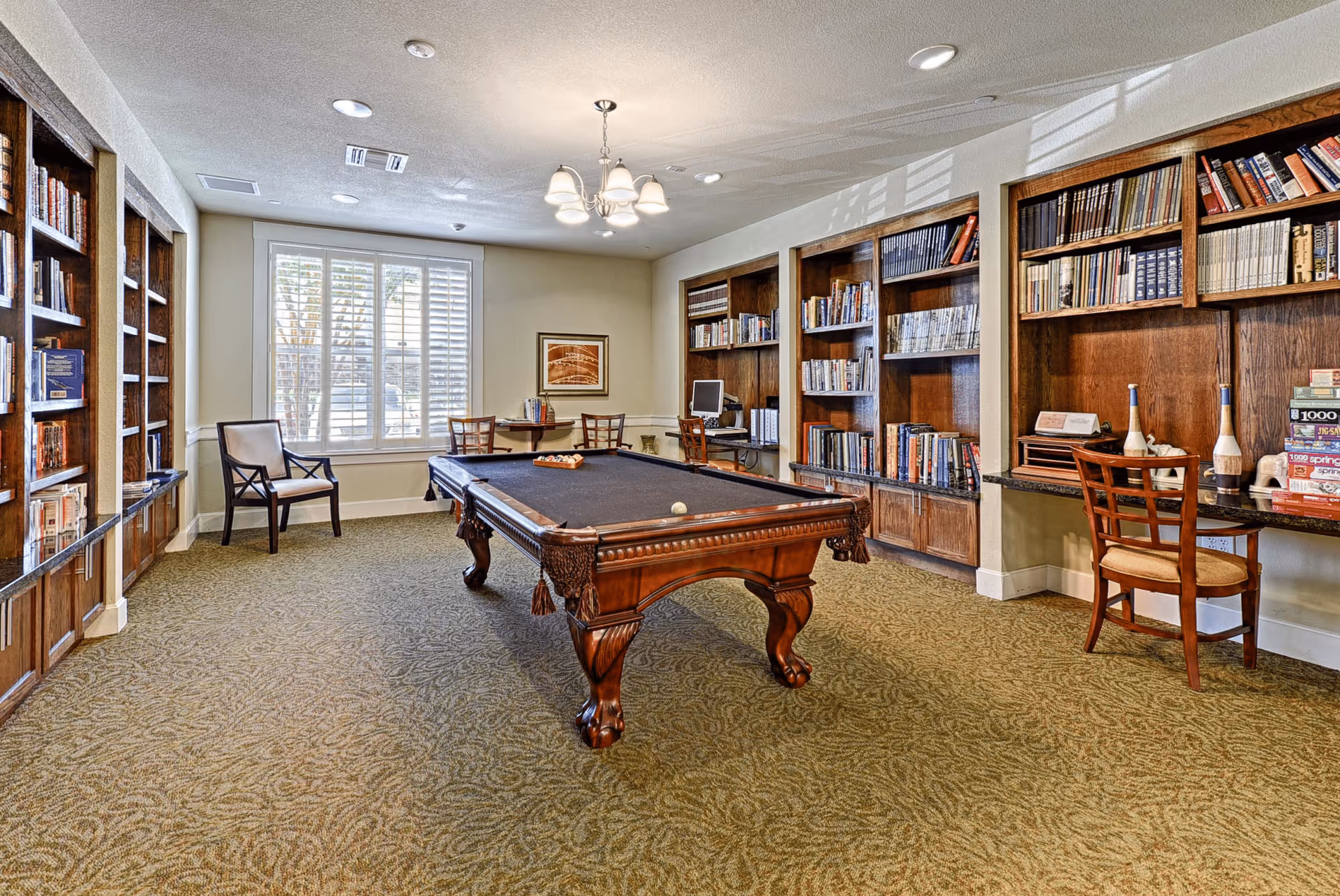 A spacious room with a pool table in the center, surrounded by built-in wooden bookshelves filled with books. There are chairs and small tables near the window with white shutters, and a computer workstation is visible against the far wall. The room has a carpeted floor and a ceiling light fixture.