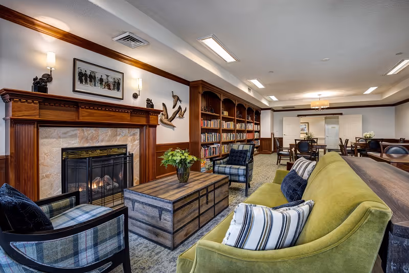 Cozy communal living room with a fireplace, built-in bookshelves, a green sofa and armchairs arranged around a wooden coffee table.