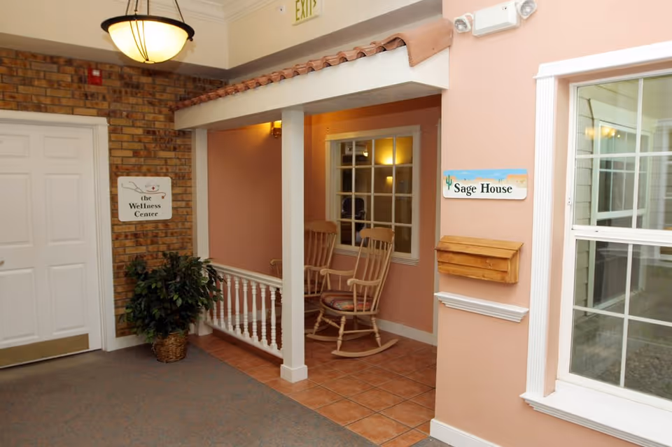 Interior hallway area in Juniper Village at Aurora featuring a small porch-like nook with two wooden rocking chairs. The walls are painted peach with white trim, and there is a window looking into another room. A sign on the brick wall reads 'the Wellness Center' and another sign on the peach wall reads 'Sage House'. A potted plant is placed near the door, and a ceiling light fixture hangs above.