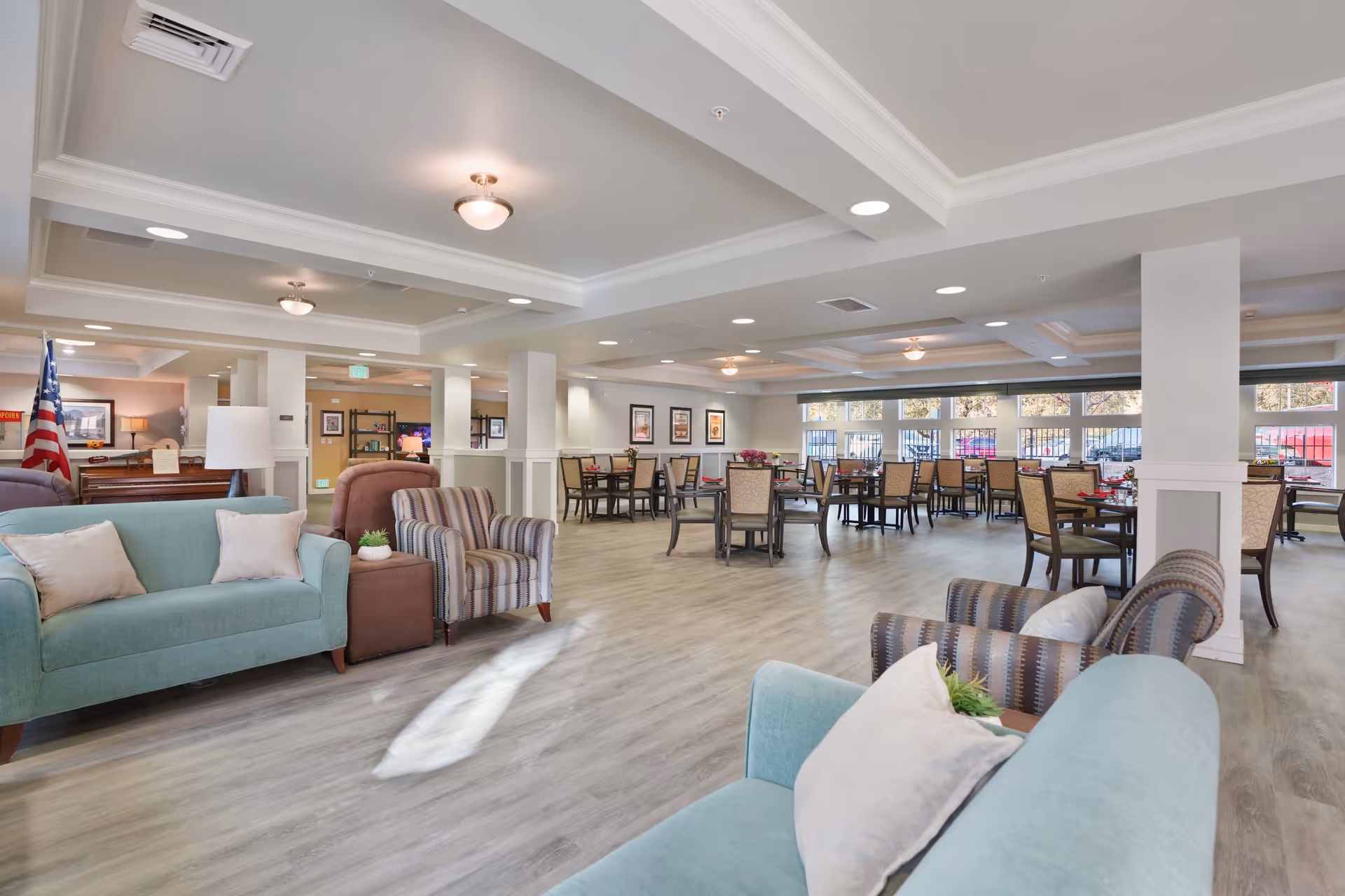 Spacious and well-lit common area in a senior living facility featuring comfortable seating with sofas and armchairs in the foreground and multiple dining tables with chairs in the background. Large windows allow natural light to fill the room, and the decor includes framed pictures on the walls and a piano with an American flag nearby.