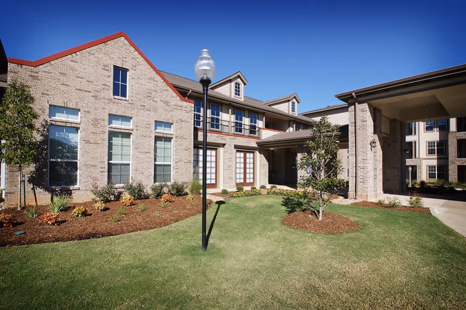 Exterior view of a senior living facility building with beige brick walls, multiple windows, a covered entrance, a well-maintained lawn, small trees, and a lamppost under a clear blue sky.